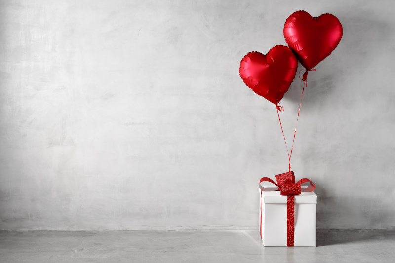 Zwei rote Herzballons und eine weiße Geschenkbox vor einer weißen Wand.